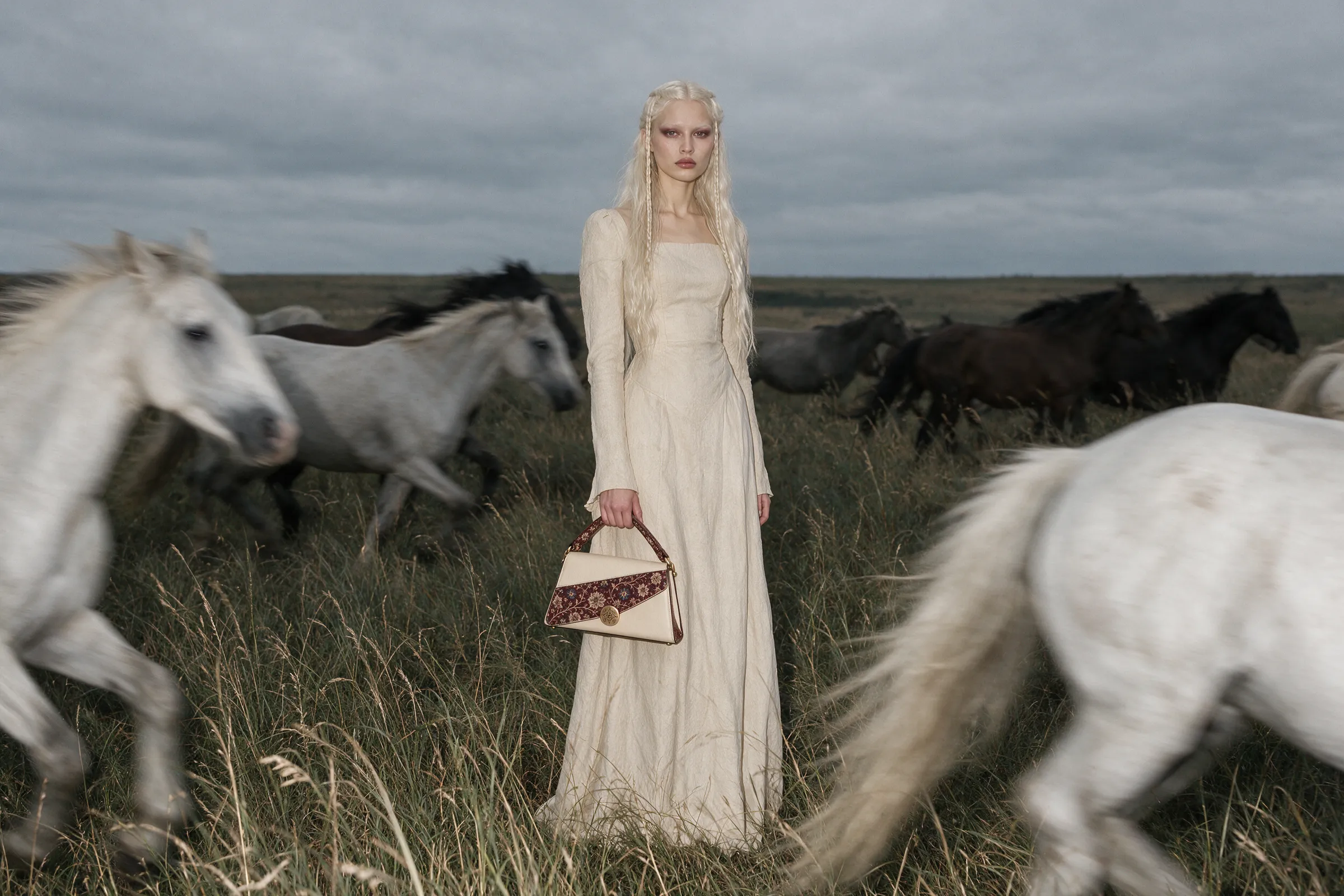 A pale model in a cream dress holding a cream and burgundy bag as white and black horses run past