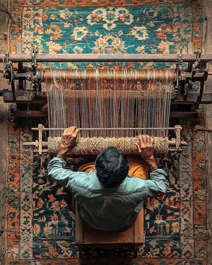A carpet weaver at a loom seen from above, working over an antique Persian carpet